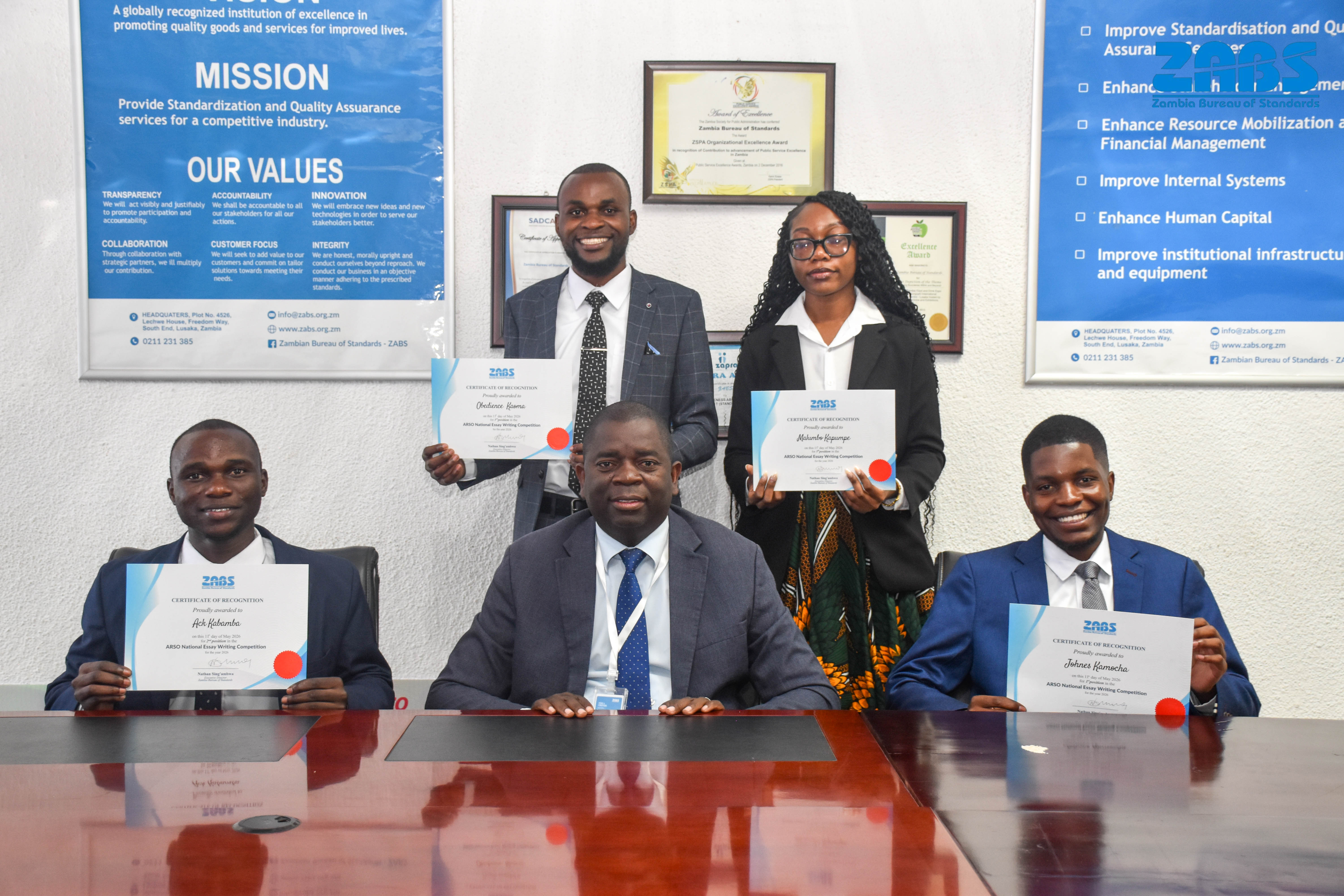 Essay competition winners and officials displaying certificates in a formal group portrait.