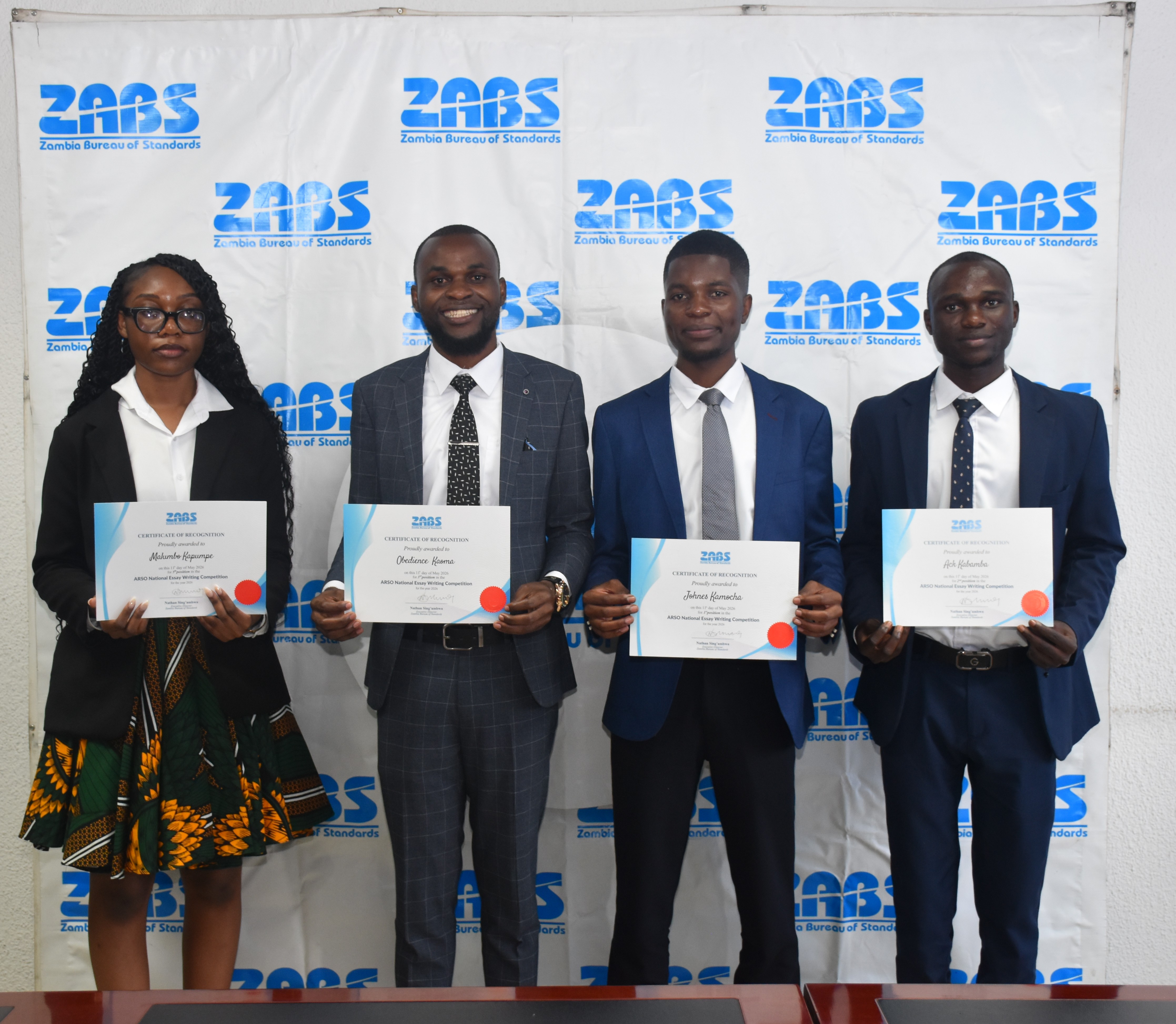 Essay competition award recipients standing with certificates at a ZABS backdrop.